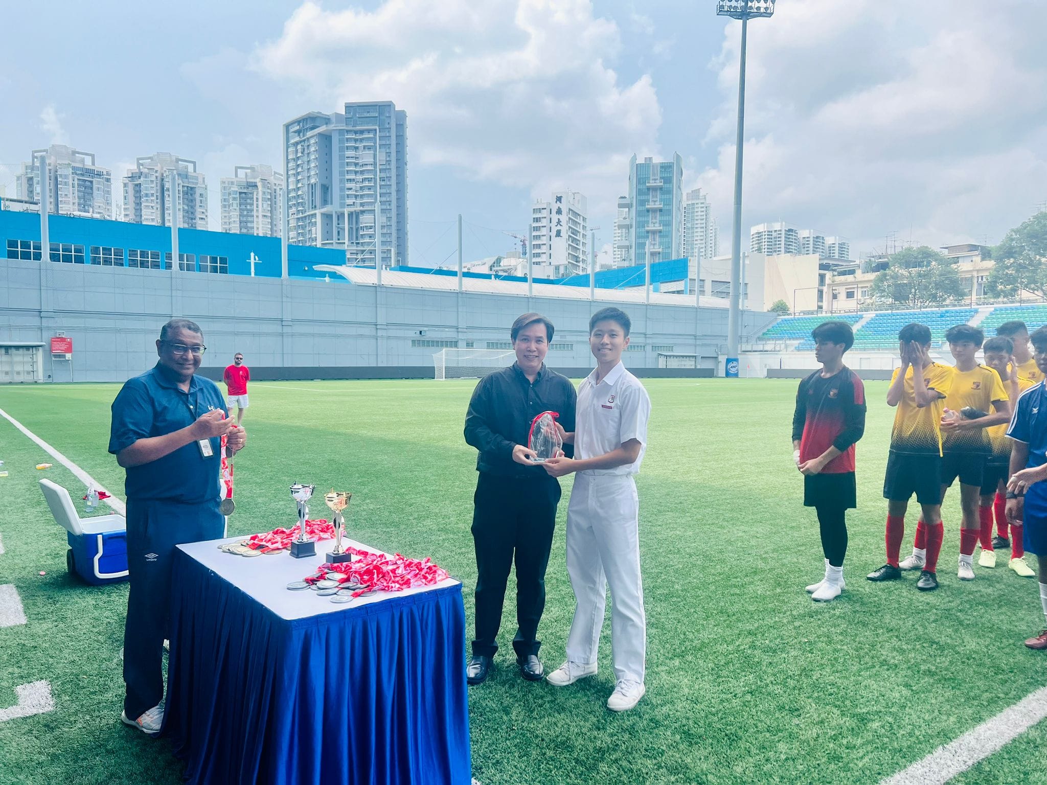 Two men present a crystal trophy on a football field, with trophies on a table and young players watching nearby.